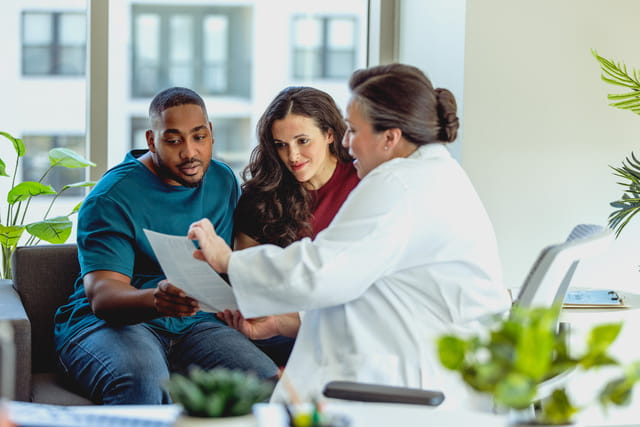 A couple sits on a sofa, attentively reviewing a document with a doctor in a white coat in a bright office with large windows and green plants.