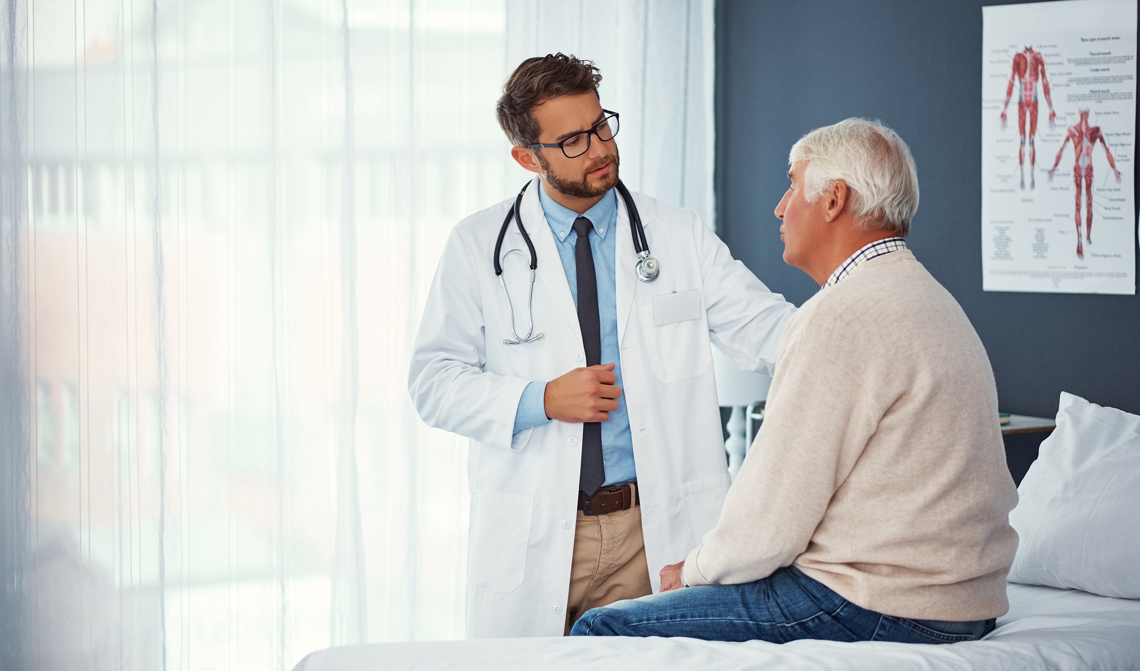 Older man sitting in doctor's office with doctor standing nearby with hand on patient's shoulder Older man sitting in doctor's office with doctor standing nearby with hand on patient's shoulder