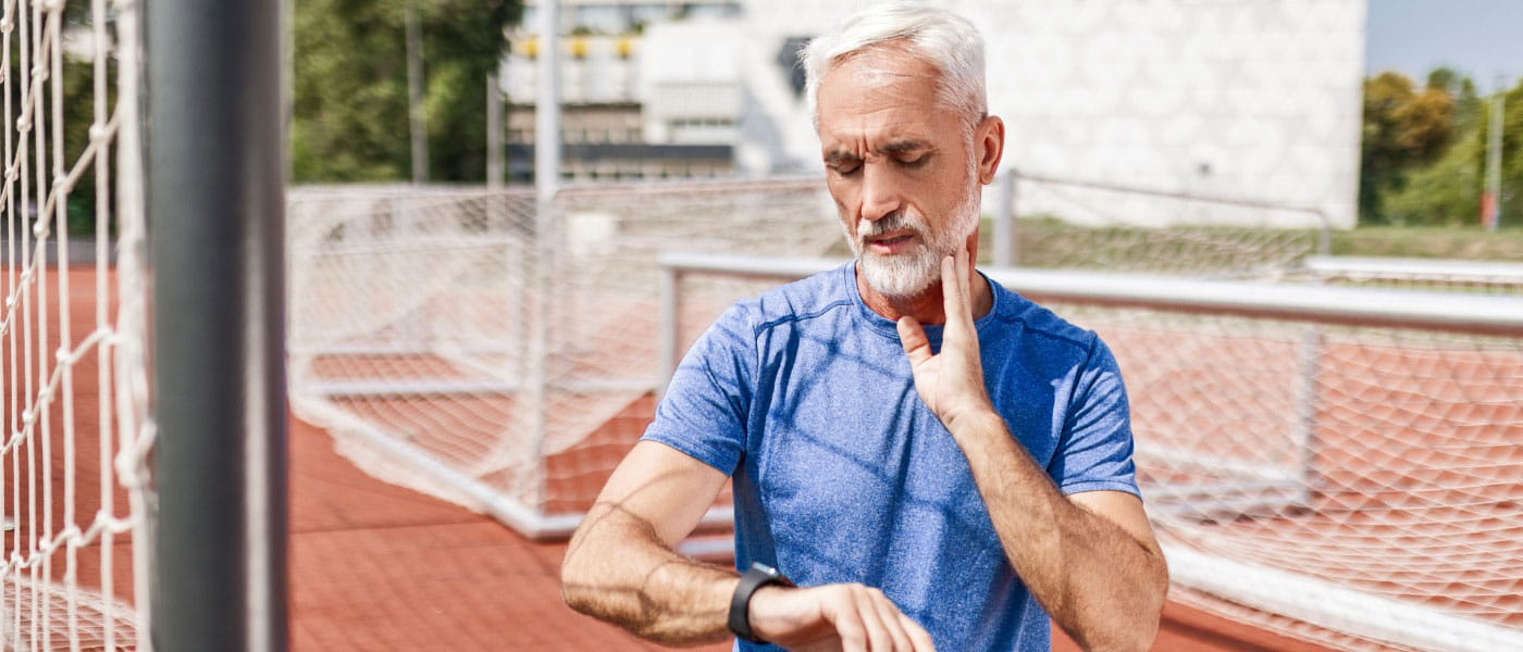 Older man touching his neck and looking at his smartwatch to measure his heart rate at an outdoor track.