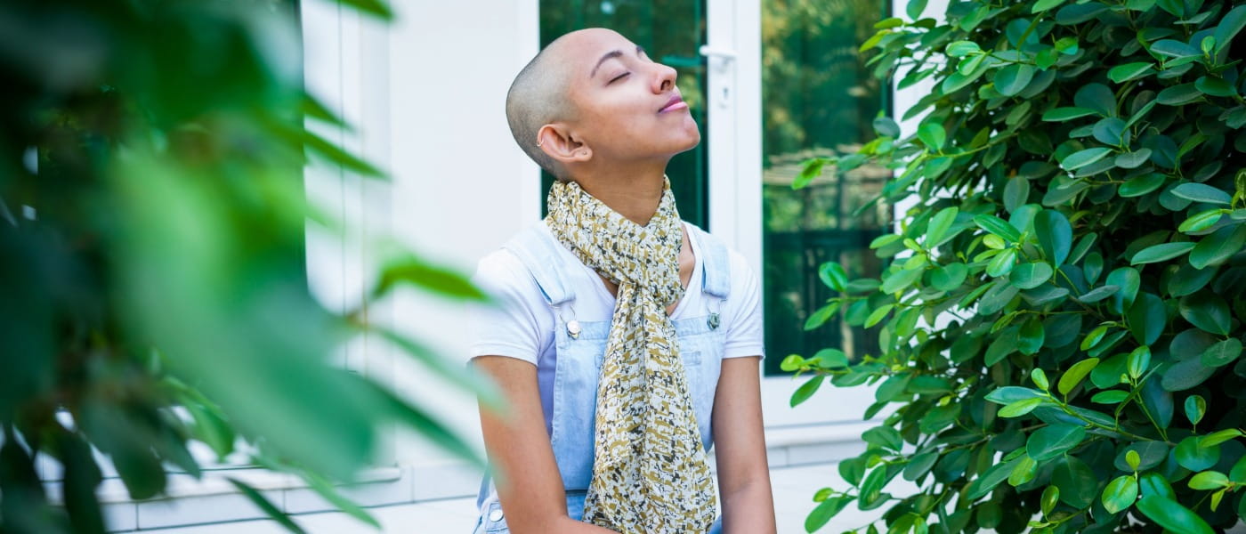 A person with a shaved head, wearing a white shirt and floral scarf, sits peacefully outdoors between green bushes, exuding tranquility.