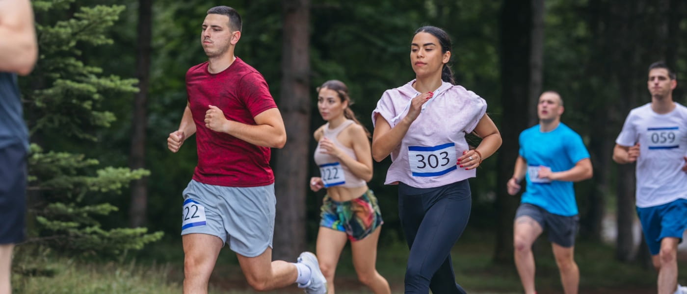 Runner leader runs ahead group of five athletes marathoners racing on a track in the woods.
