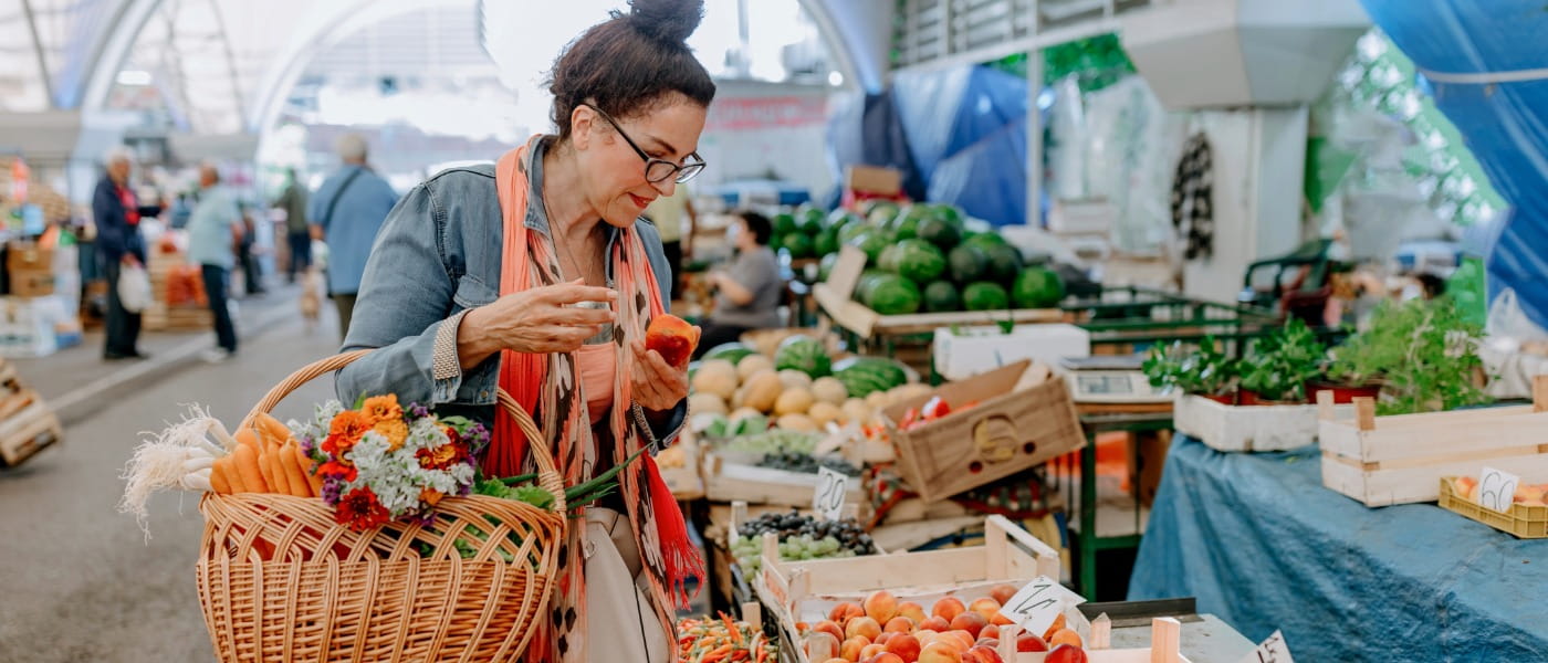 A woman holding a pepper and examining a crate full of peaches inside a farmers' market, while carrying a basket filled with fresh flowers and vegetables.
