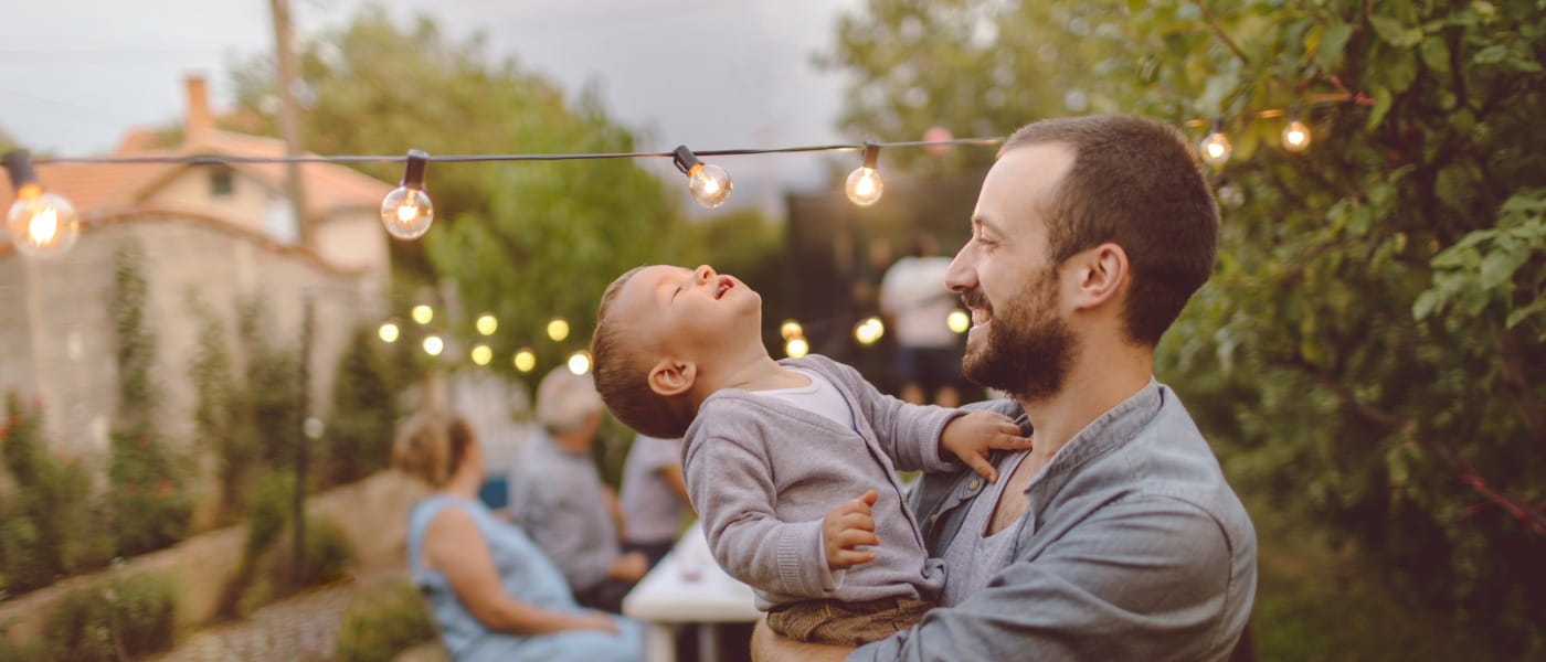 Young father holding his toddler son in his arms and both are smiling in front of their multi-generation family having dinner outdoors in their back yard.