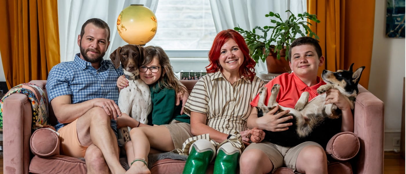 Kaitlin sitting on the family couch in between her children Declan and Finlay along her husband Matt and two dogs.