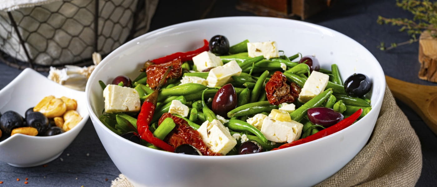 White bowl of green bean salad with olives, feta cheese and sun-dried tomatoes set on a table next to a bowl of berries and basket of green beans.