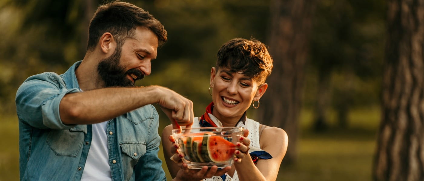 Young man and woman sharing a bowl of watermelon slices during a picnic in a wooded park.