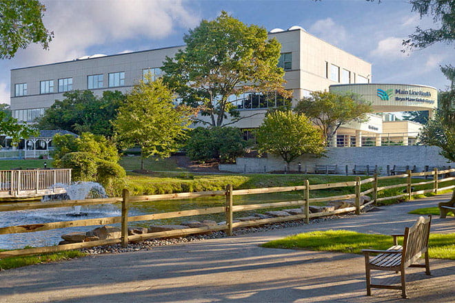 Bryn Mawr Rehab Hospital view from outside, with the main entrance on the right. A pond with a fountain and a surrounding brown fence sits in front of the building. The sky is blue behind the building.